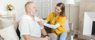 A nurse uses a stethoscope for a home check-up on a senior adult in a cozy living room.